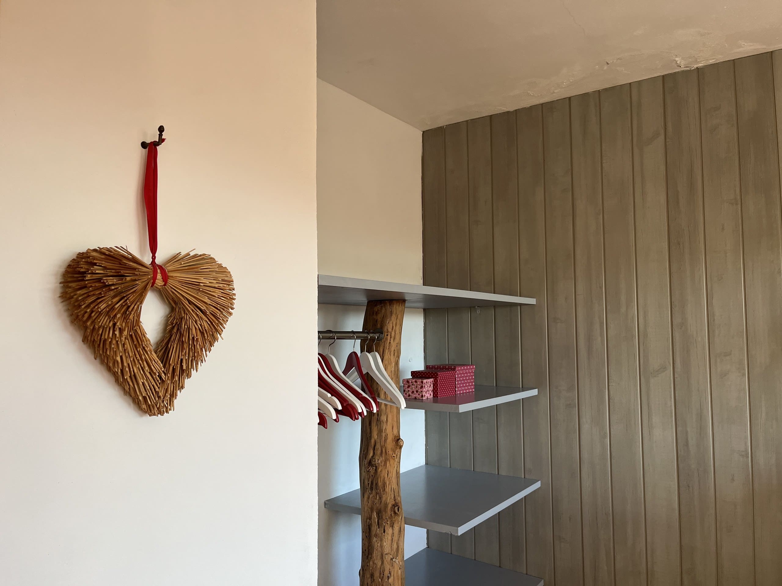 Offene Kleiderablage mit Regalbrettern und Bügeln in der Casa Amor Perfeito, daneben ein Strohherz an der Wand.