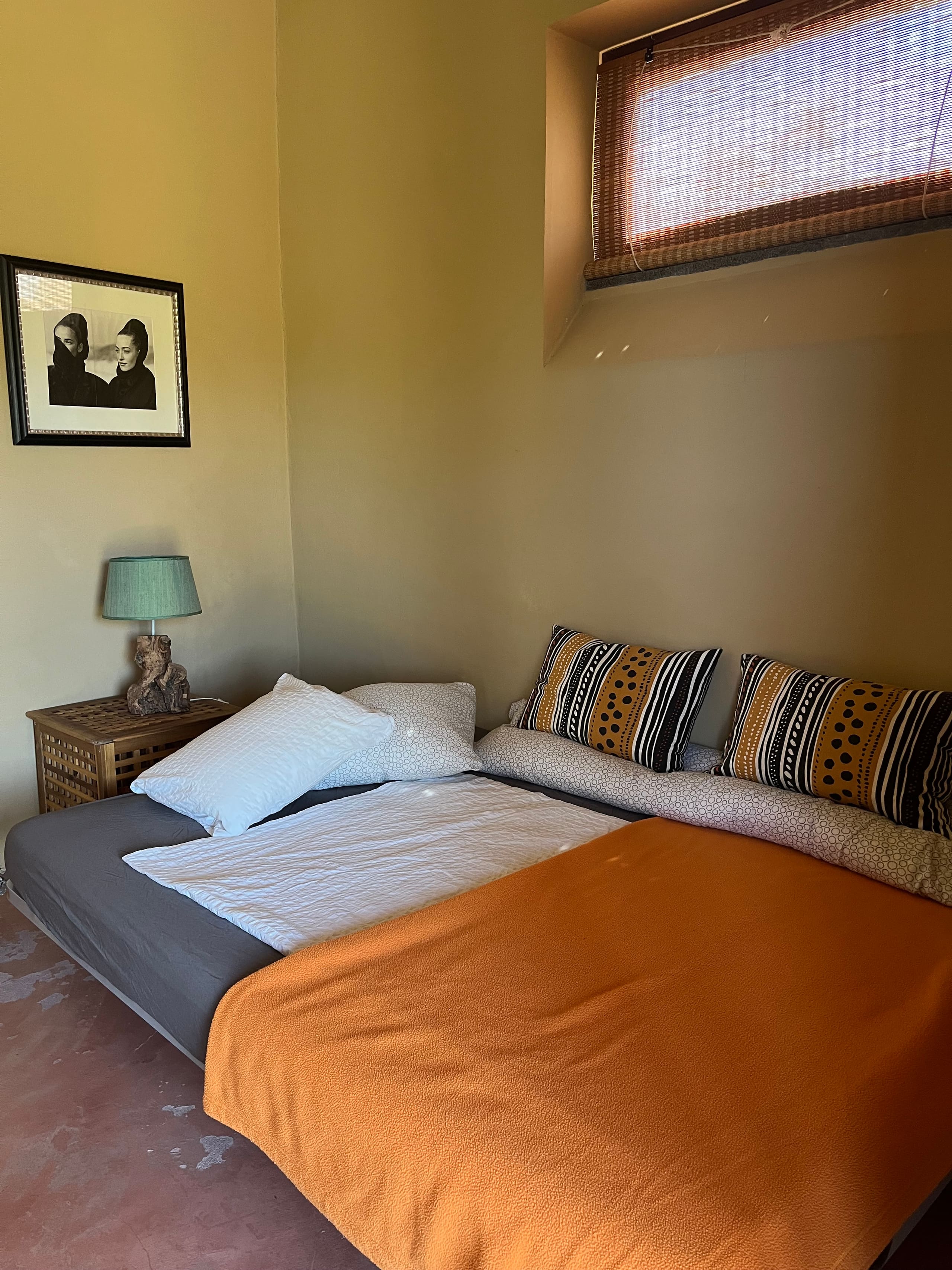 Bett mit oranger Tagesdecke und gemusterten Kissen unter dem kleinen Fenster der Casa Terra.