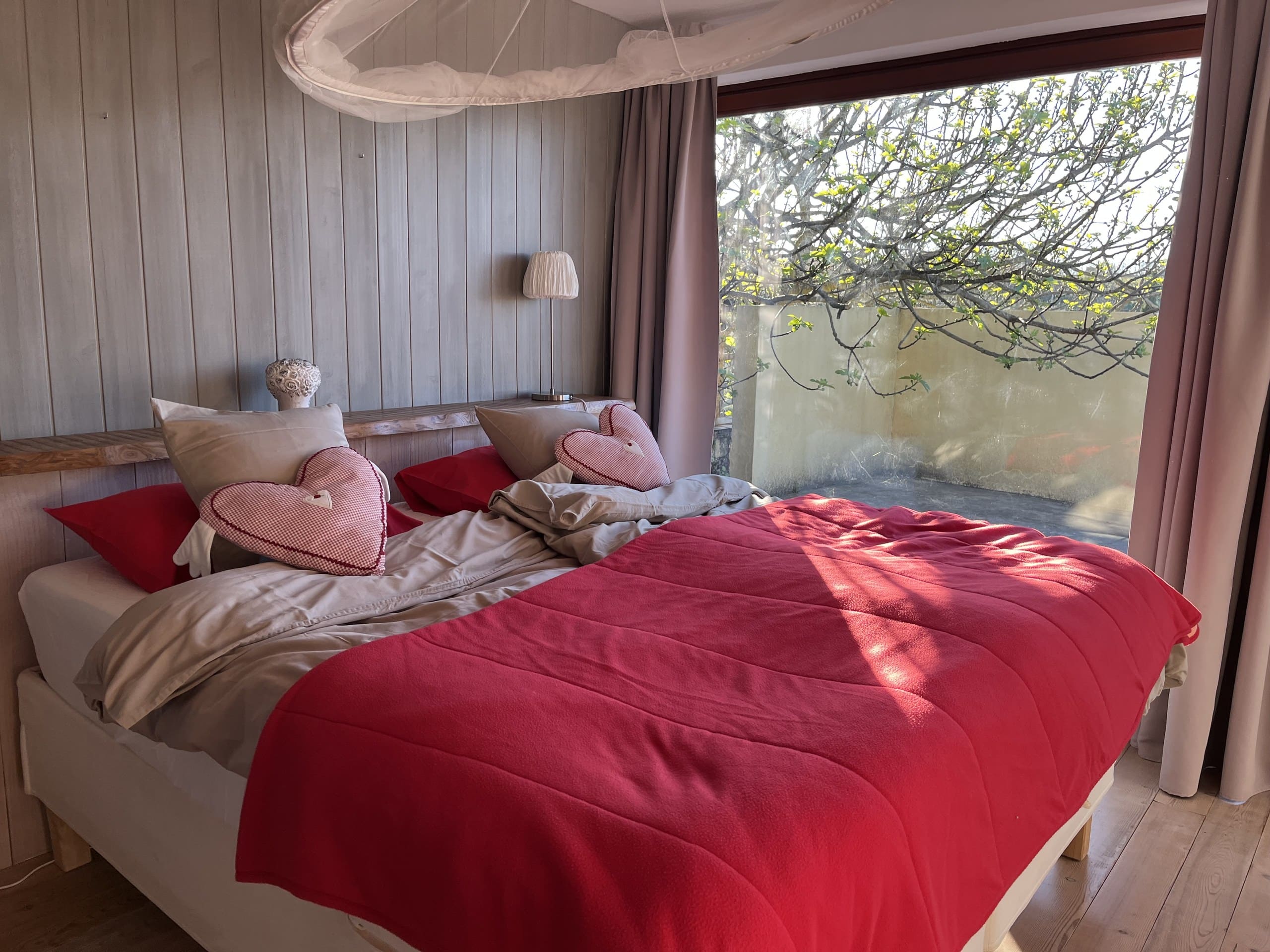 Doppelbett mit roter Decke und Herzkissen in der Casa Amor Perfeito, dahinter der Feigenbaum vor dem Fenster.