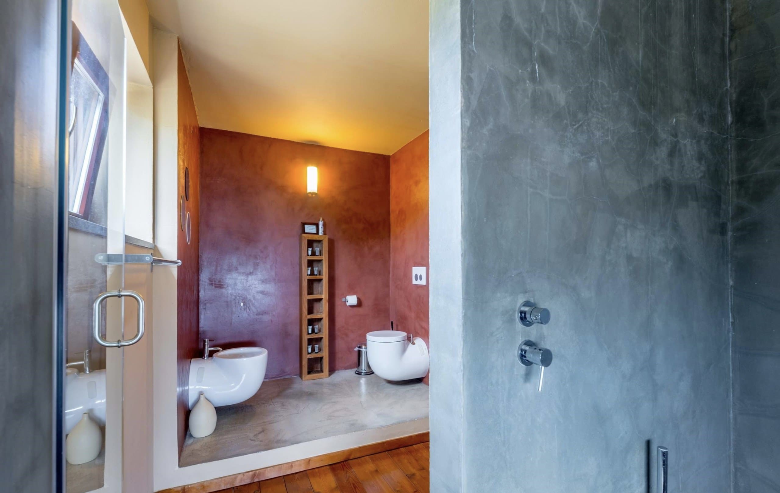 Dusche der Casa Terra mit grauer Putzwand, dahinter der Blick in den roten Sanitärbereich.