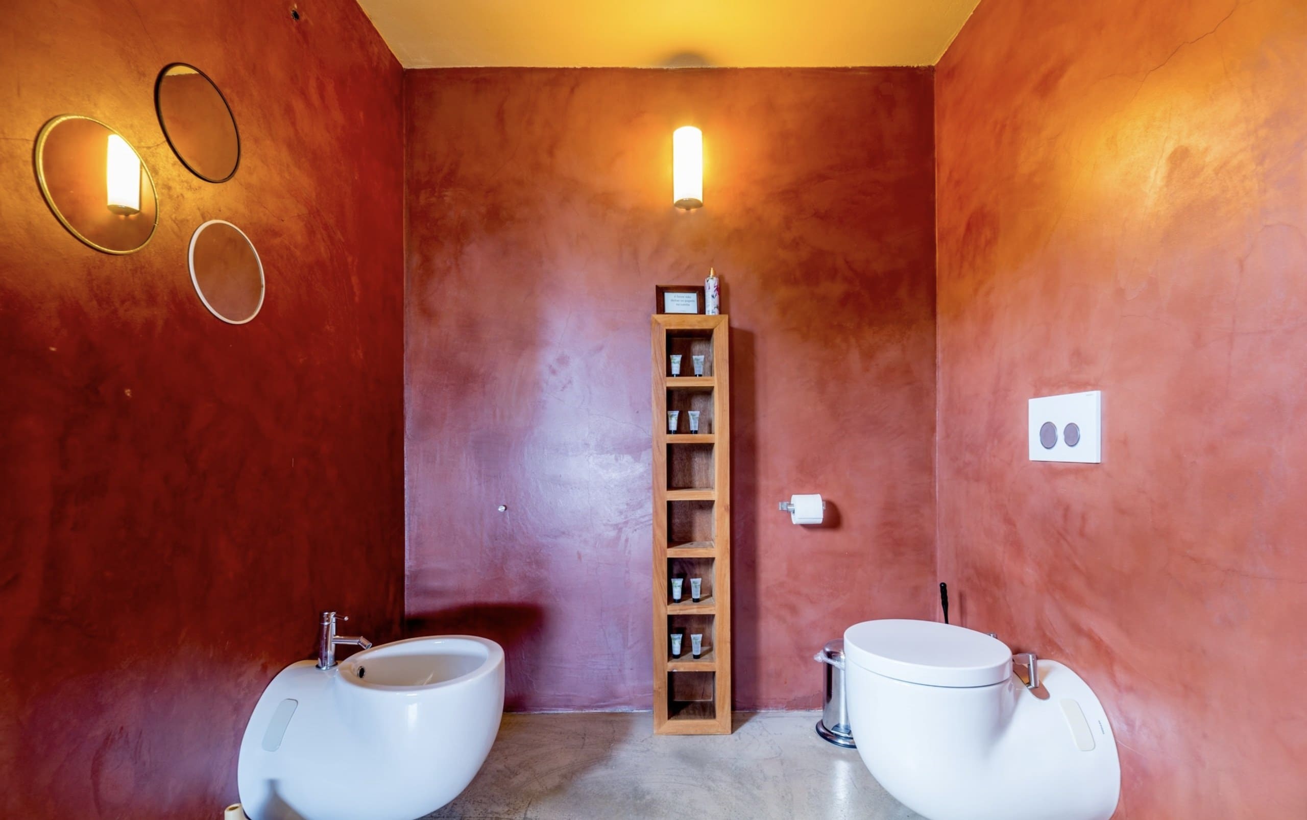 Bad der Casa Terra mit terrakottaroten Wänden, WC, Bidet und schmalem Holzregal.