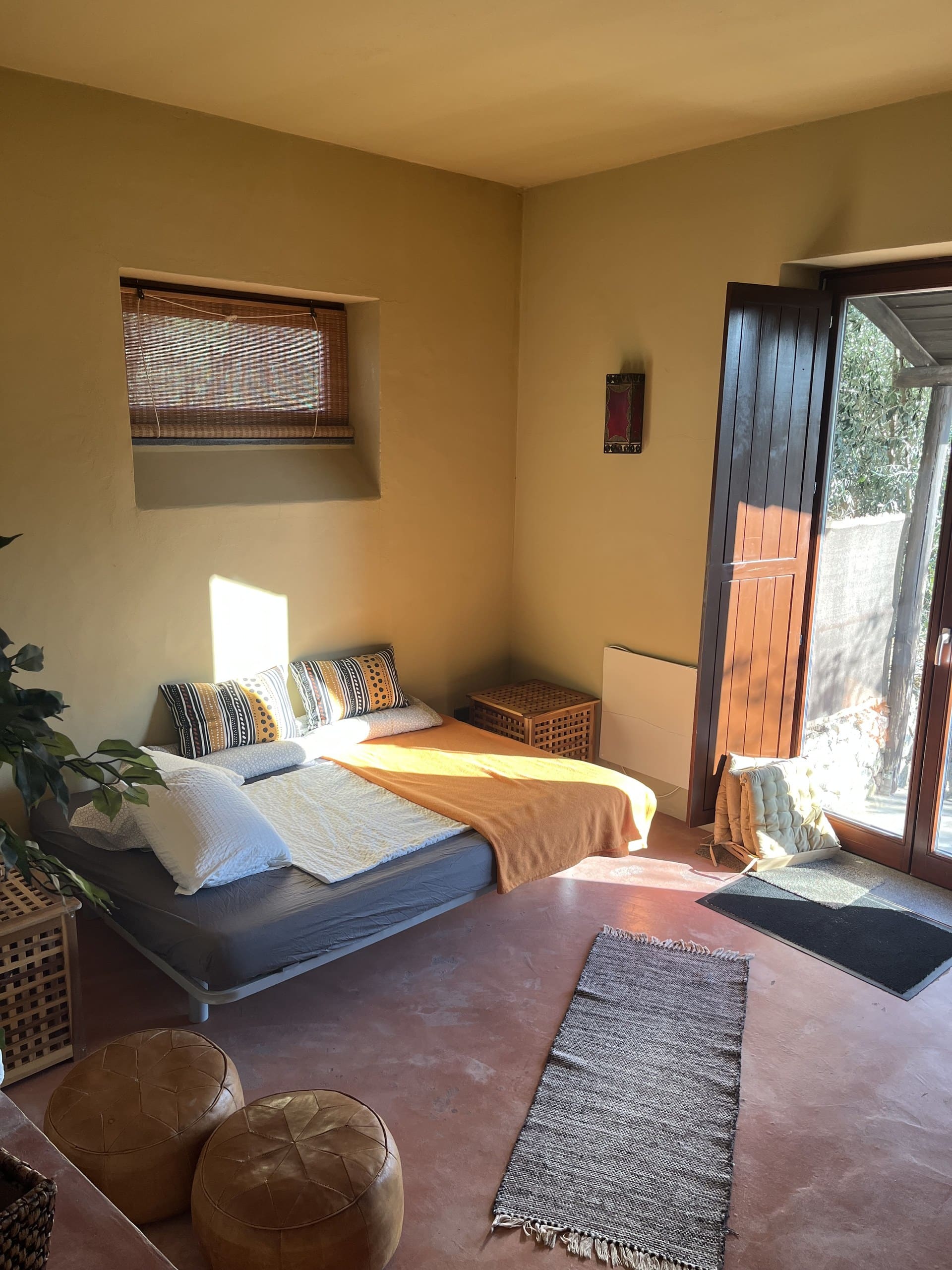 Zweites Bett im Wohnraum der Casa Terra, daneben die Fenstertür in den Garten und zwei Lederpoufs.