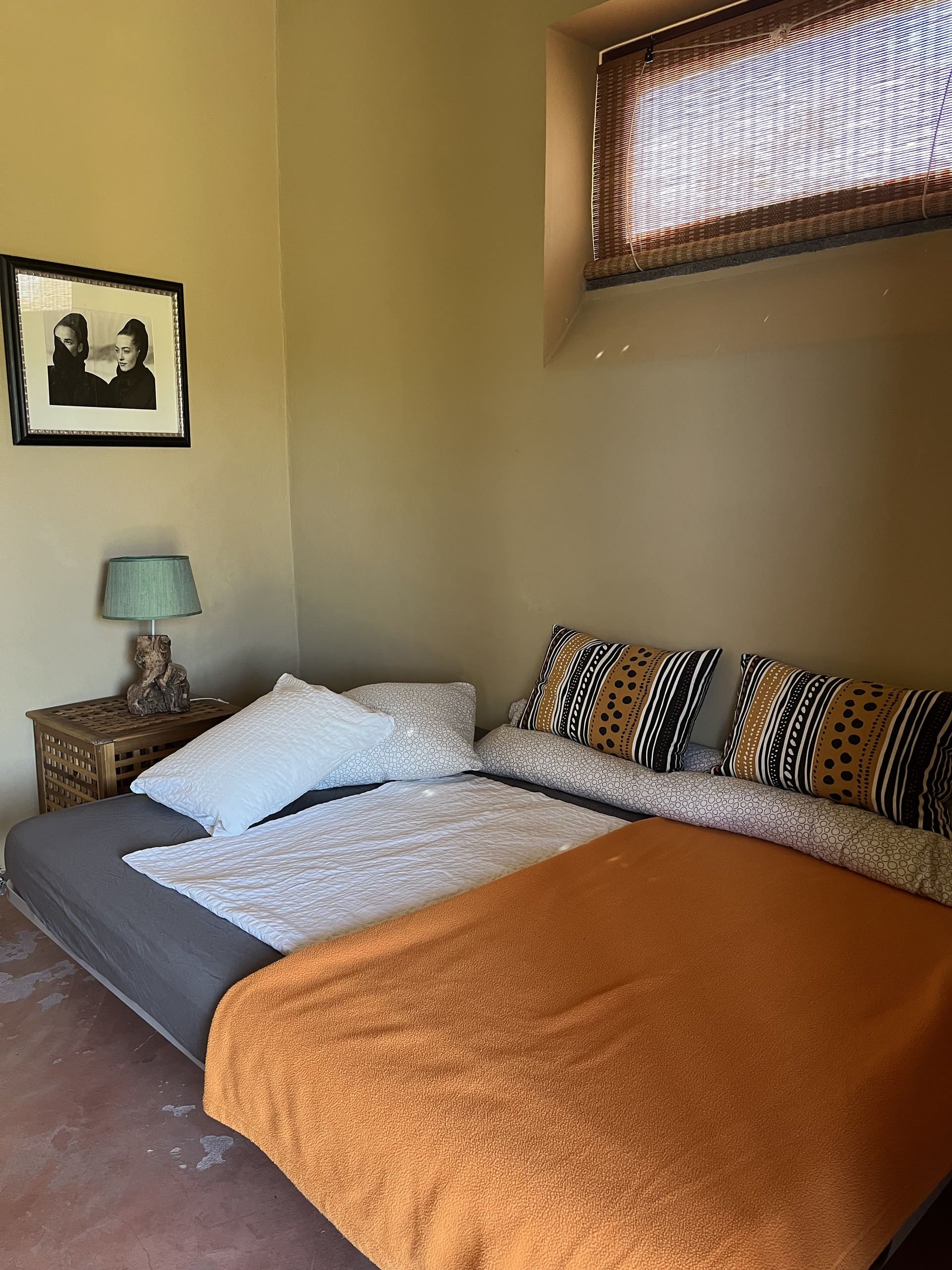 Bett mit oranger Tagesdecke und gemusterten Kissen unter dem kleinen Fenster der Casa Terra.