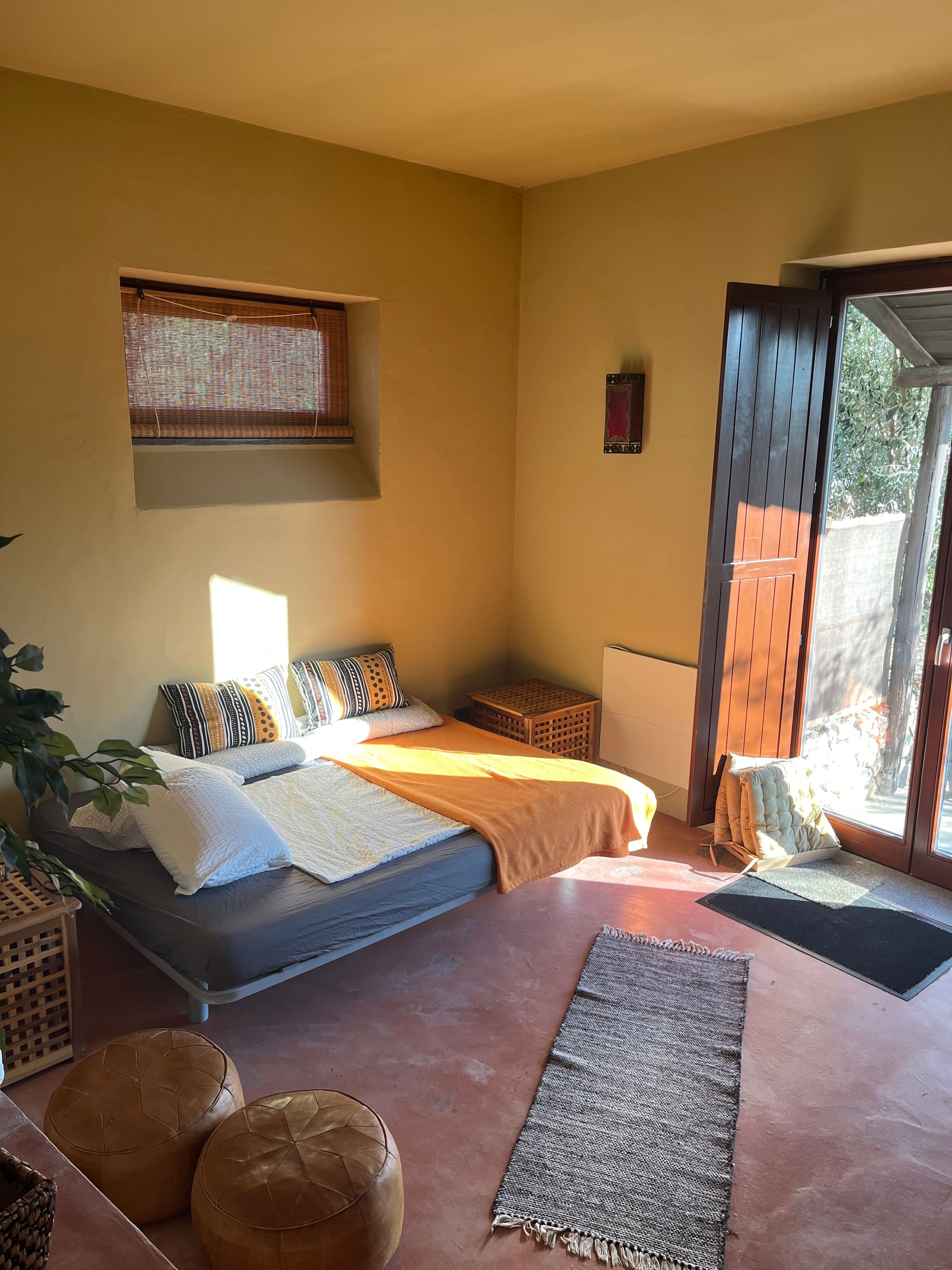 Zweites Bett im Wohnraum der Casa Terra, daneben die Fenstertür in den Garten und zwei Lederpoufs.
