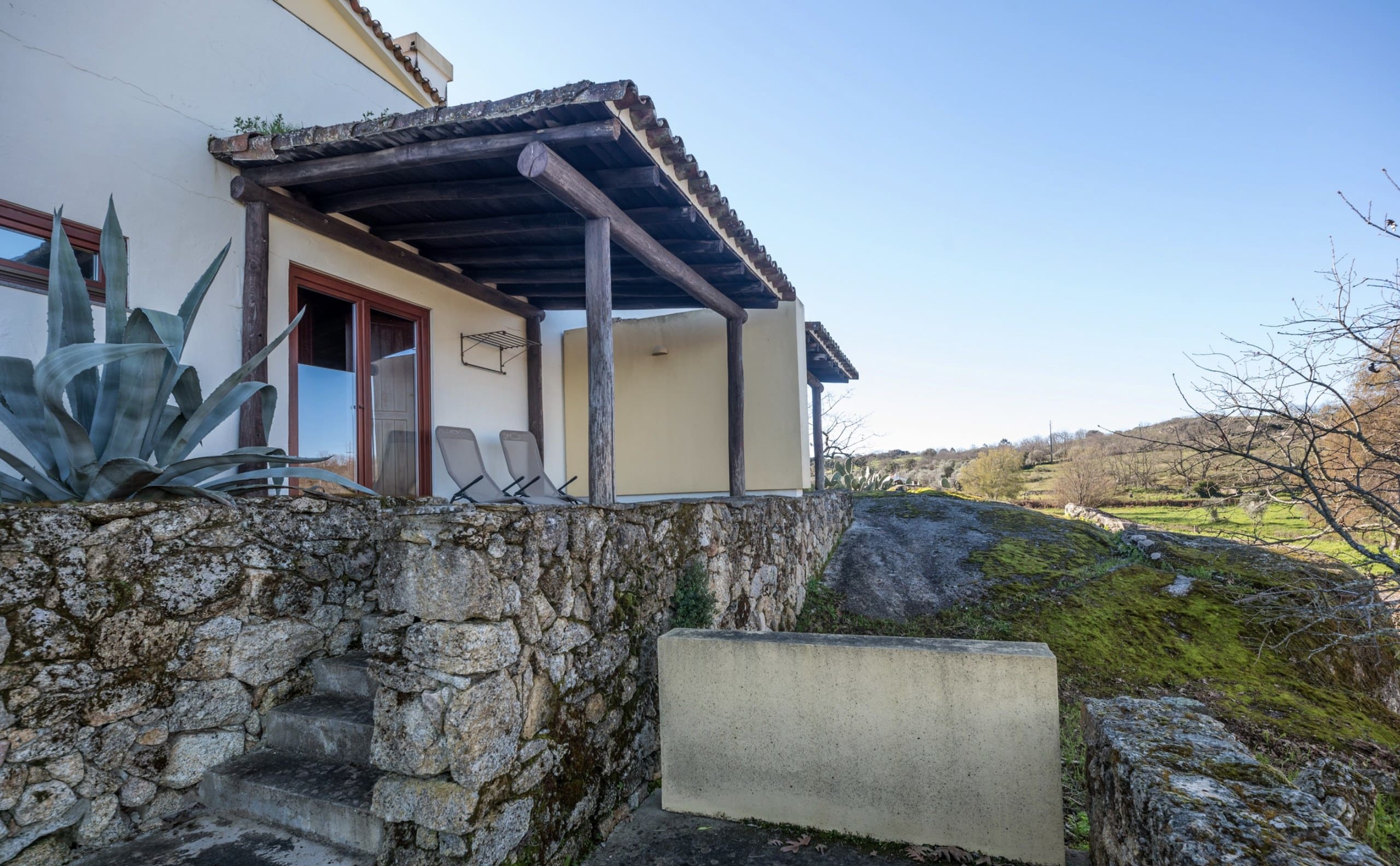 Außenansicht der Casa Hridaya mit überdachter Terrasse über einer Granitmauer.