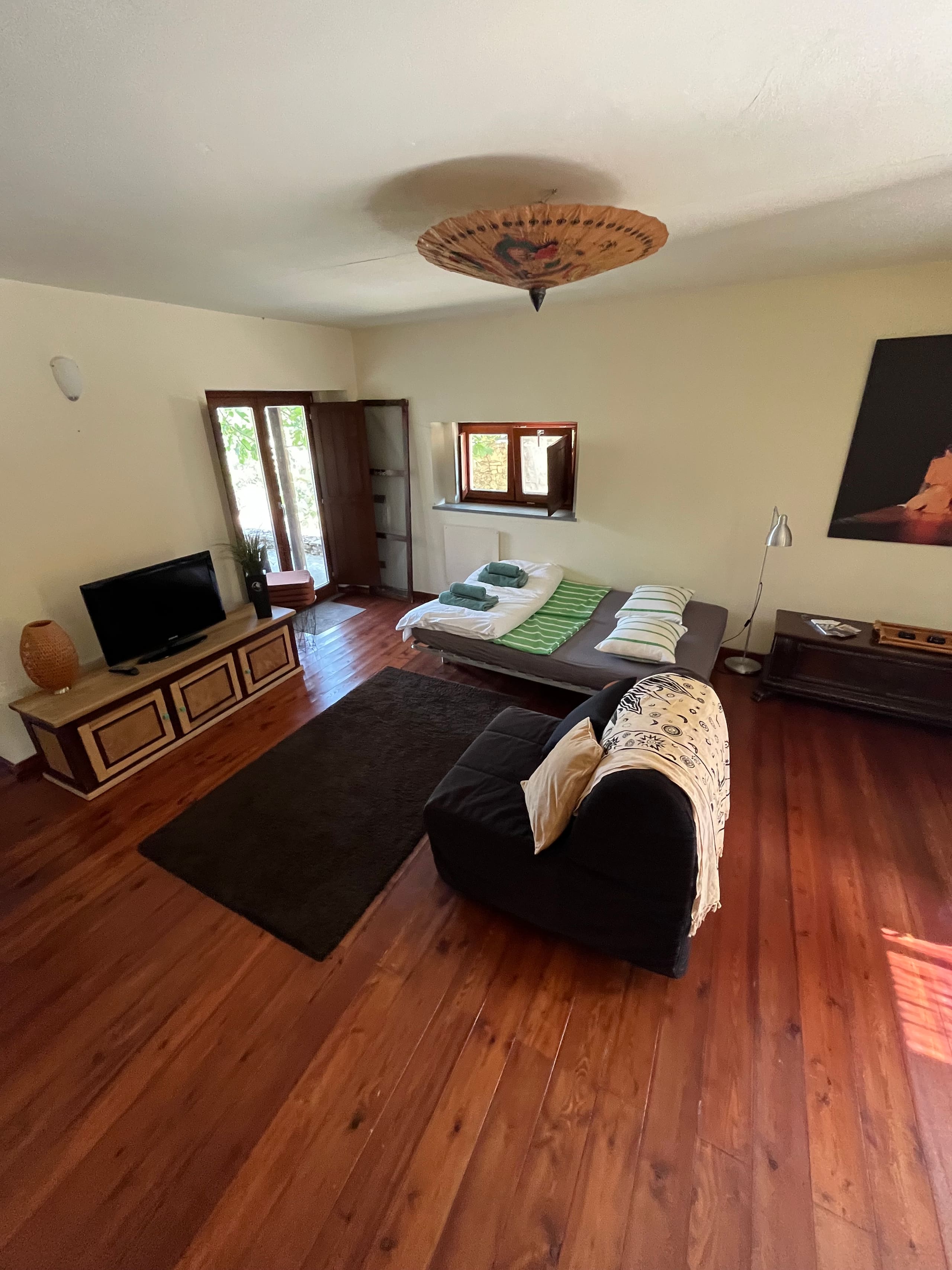 Wohnraum der Casa Hridaya mit Fernsehkommode, Tagesbett und Sessel auf Holzdielen.