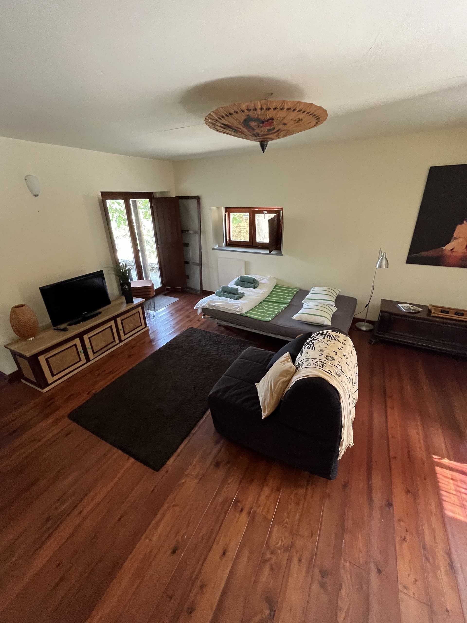 Wohnraum der Casa Hridaya mit Fernsehkommode, Tagesbett und Sessel auf Holzdielen.